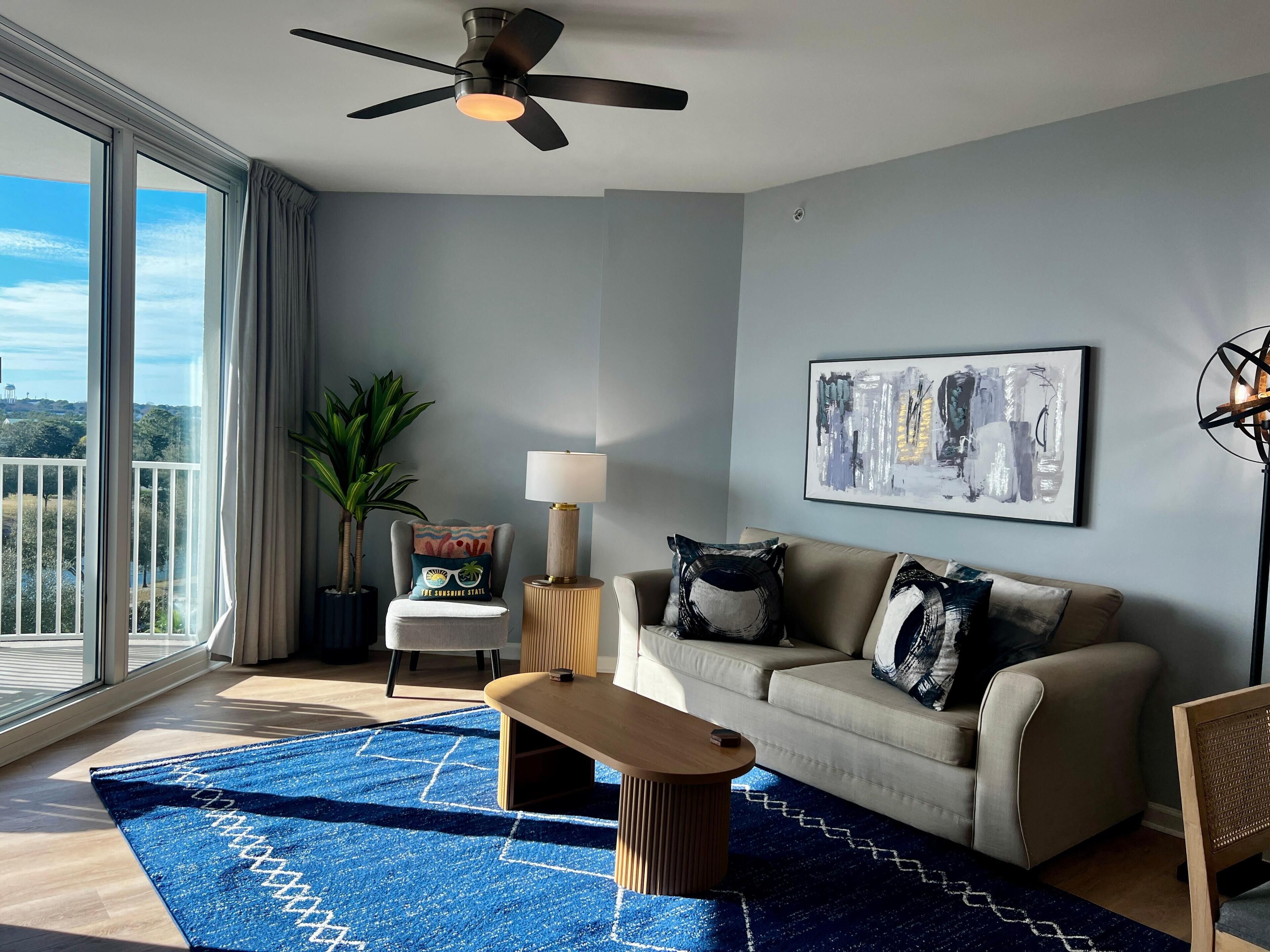 Un salon moderne doté d’une grande fenêtre, d’un ventilateur de plafond, d’un canapé, d’une table basse et d’un lampadaire.