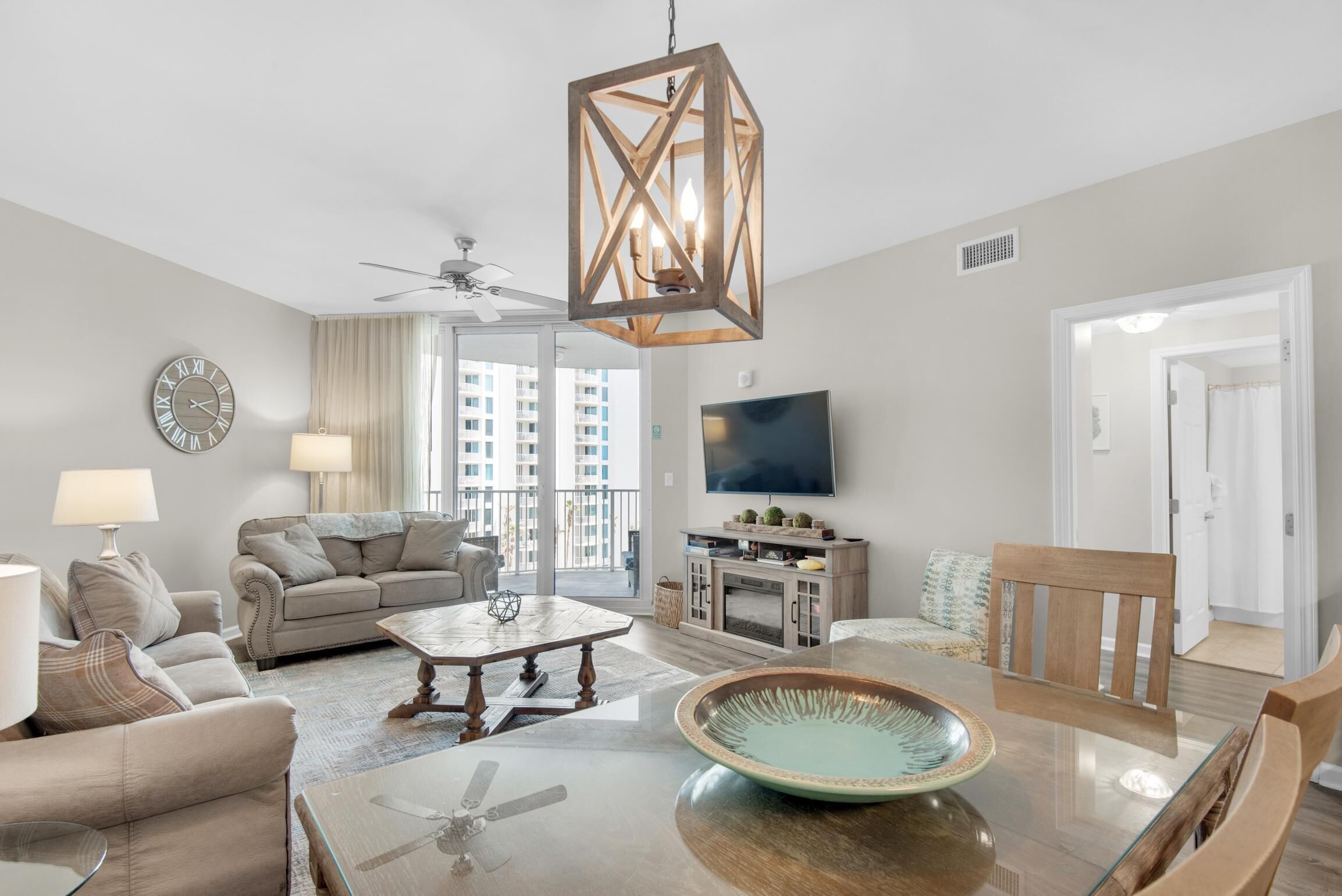 A spacious living room with a ceiling fan, a wooden dining table with a glass top, a large mirror, and a television on the wall.