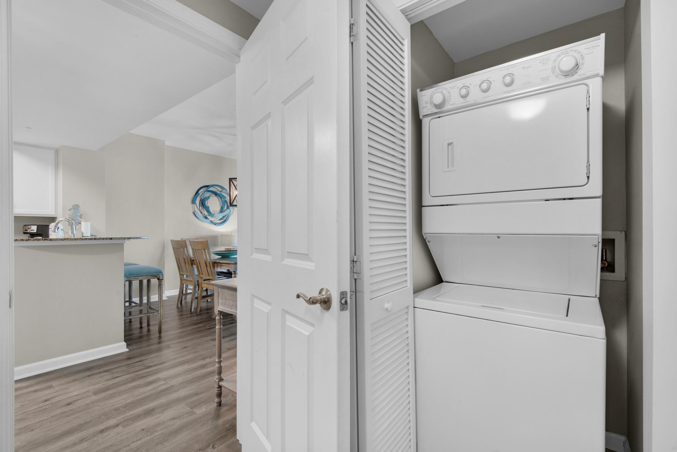 Uma lavanderia com máquinas de lavar e secar empilhadas, uma porta que dá para uma sala de jantar e uma cozinha ao fundo.