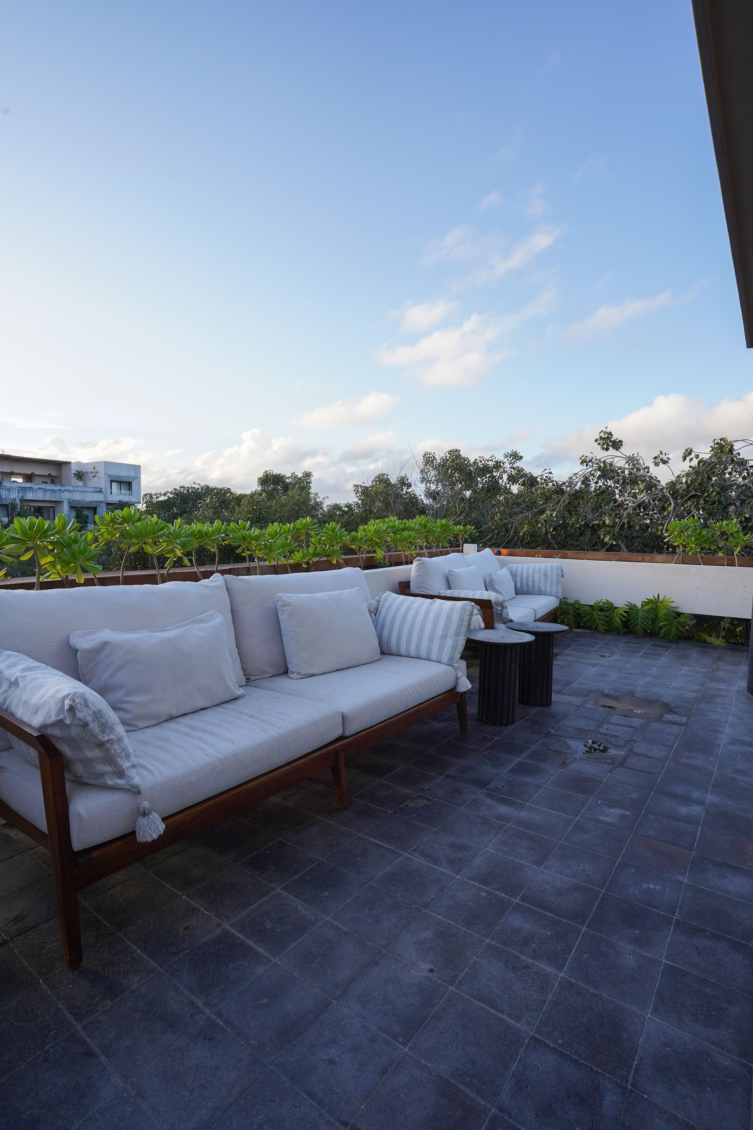 A rooftop patio with a sofa and round tables, surrounded by greenery and a clear sky.