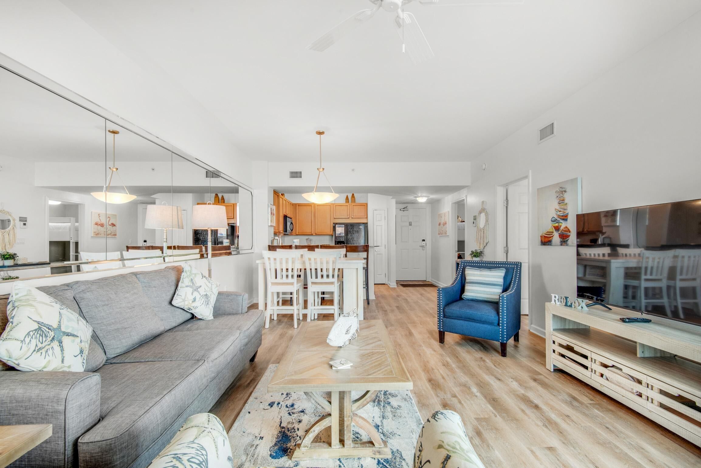 A modern living room with a grey sofa, a blue armchair, and a wooden coffee table. There is a kitchen in the background with wooden cabinets and a white countertop.