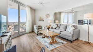 A spacious living room with a large window, a sofa, a coffee table, and a TV. - Palms Resort #21204 Jr. 2BR (Destin)