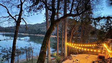 A lakeside cabin with a deck, surrounded by tall trees and a view of the lake and mountains.