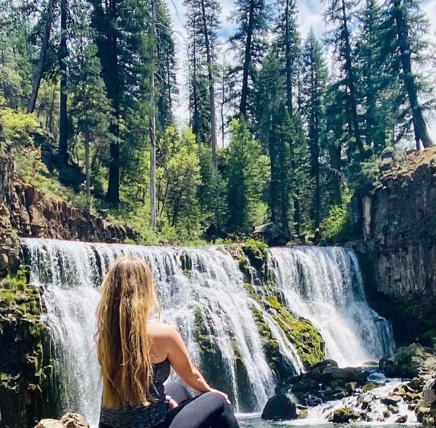 A waterfall in a forested area with a person sitting in front of it.