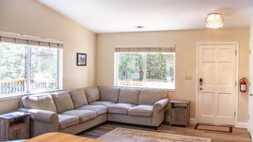 Ein Wohnzimmer mit einem beigefarbenen Ecksofa, einem Holztisch und einem Teppich. Zwei Fenster mit Jalousien, eine Tür mit Fußmatte und ein kleiner Beistelltisch sind ebenfalls zu sehen.