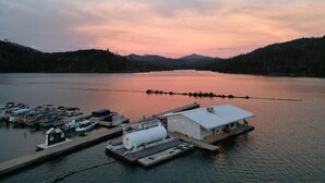 Una marina amb vaixells amarrats, un edifici blanc amb teulada plana i un llac tranquil amb muntanyes al fons.