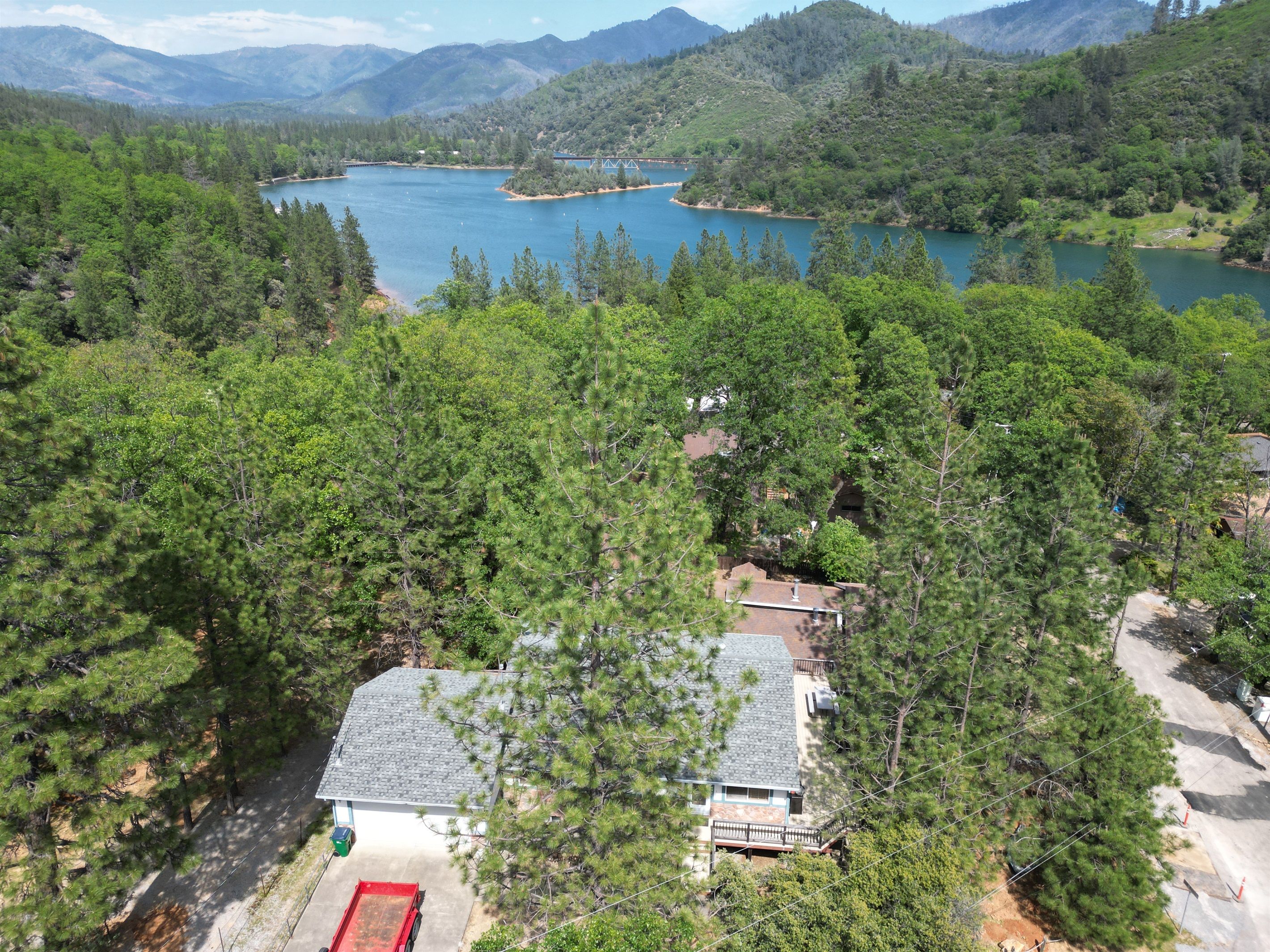 A residential area with a house surrounded by dense forest, a clear view of a lake, and a bridge in the distance.
