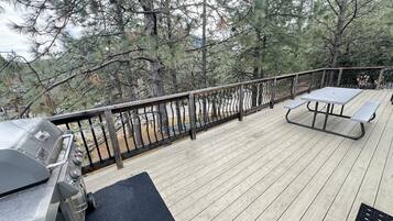 A deck with a grill, picnic table, and benches, surrounded by trees.