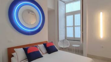 A modern hotel room with a bed, a circular blue light fixture, a window with curtains, and a small table with a chair.