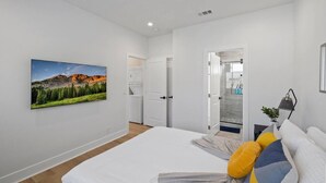 A modern bedroom with a large bed, a painting of a mountain landscape, a walk-in closet, and a bathroom visible through an open door. - Cozy Prvate ADU close to Disney Beach EV (Garden Grove)