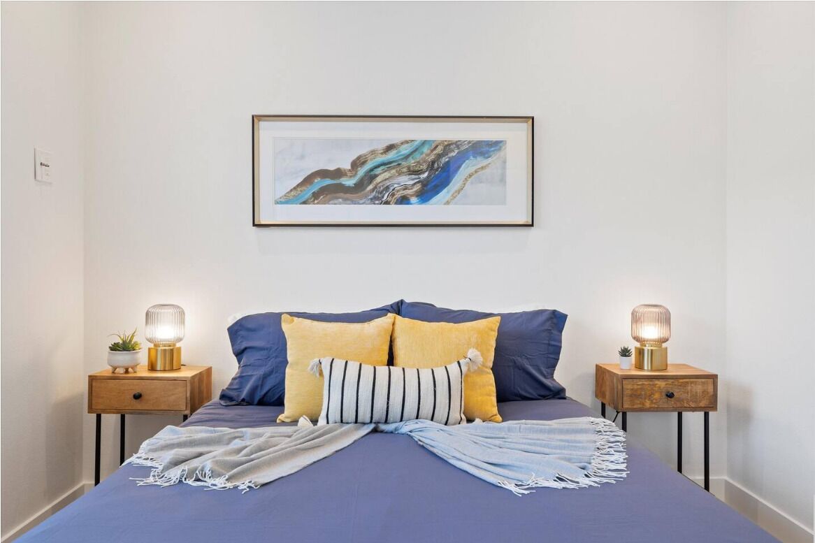 Un dormitorio con una cama, dos mesitas de noche, un cuadro abstracto enmarcado y una mesita de noche con una lámpara.
