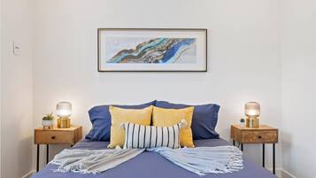Un dormitorio con una cama, dos mesitas de noche, un cuadro abstracto enmarcado y una mesita de noche con una lámpara.