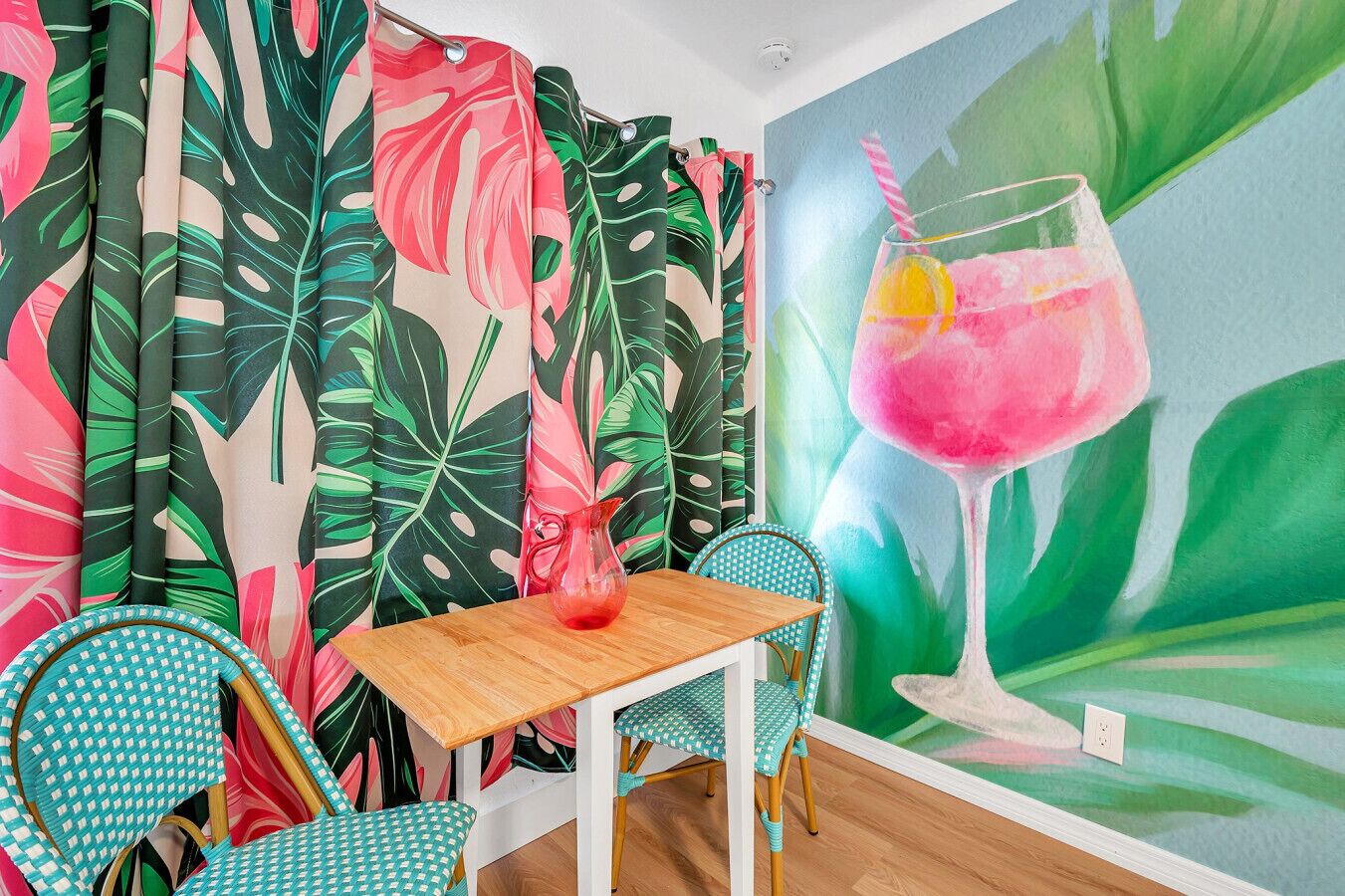 A room with tropical-themed curtains, a mural of a drink, and a wooden table with chairs.