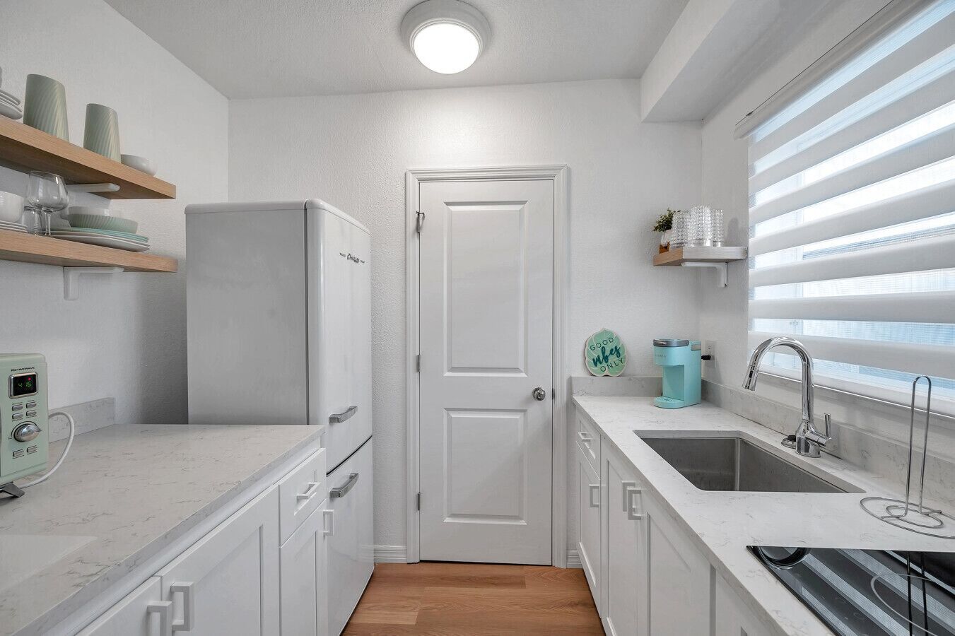 A modern kitchen with white cabinetry, a built-in refrigerator, a coffee machine, and a sink with a window above.