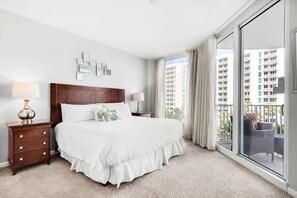 A hotel room with a large bed, a wooden nightstand with a lamp, a wall-mounted light fixture, and a balcony with a view of high-rise buildings.