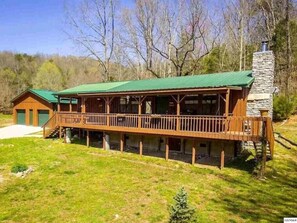 A wooden cabin with a green roof, a stone chimney, and a covered deck.