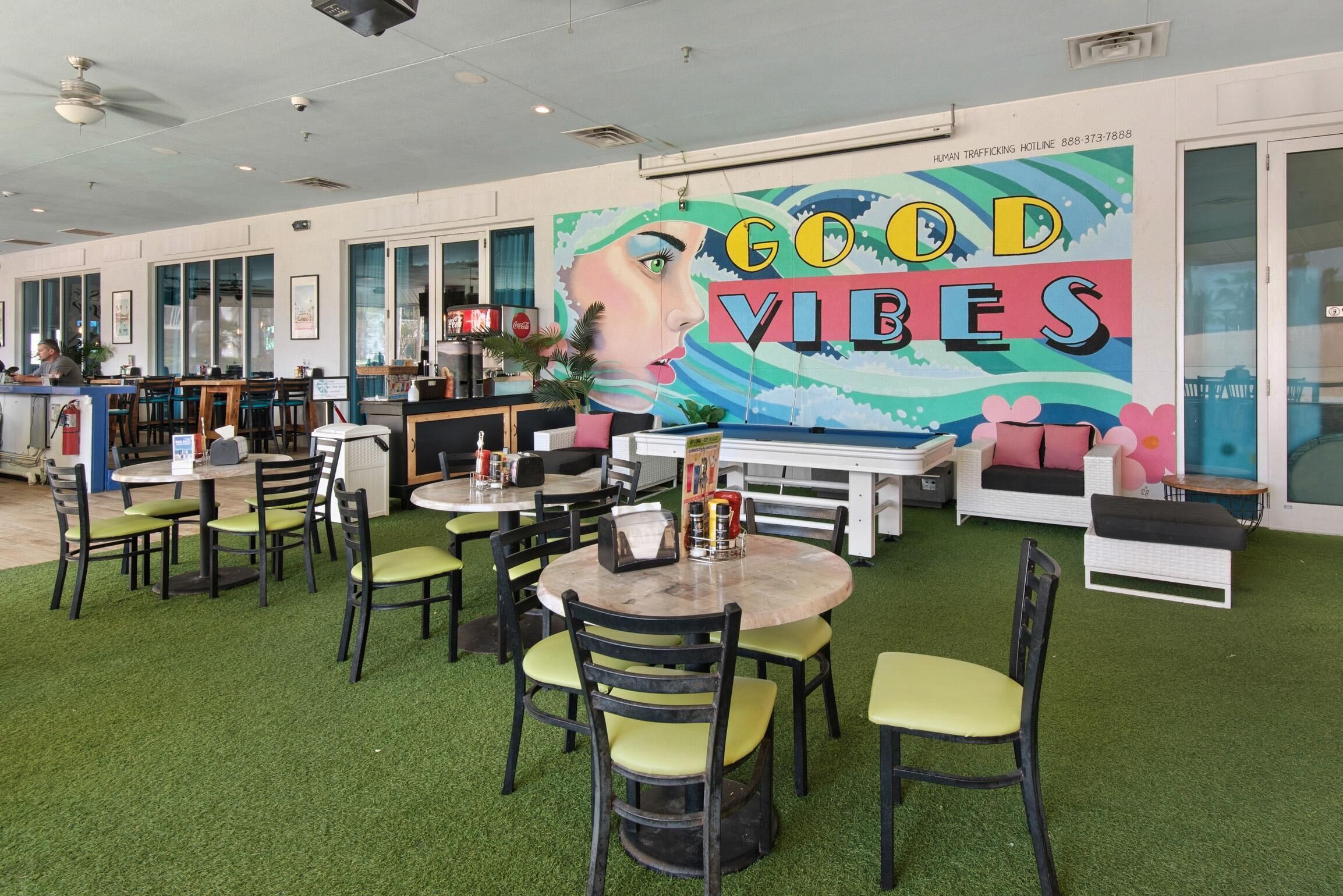 Un restaurant avec un sol recouvert de gazon artificiel vert, des tables rondes et des chaises. On y trouve une fresque murale avec le texte « BONNE AMBIANCE » et un numéro de téléphone pour une ligne d’assistance téléphonique concernant la traite des êtres humains.