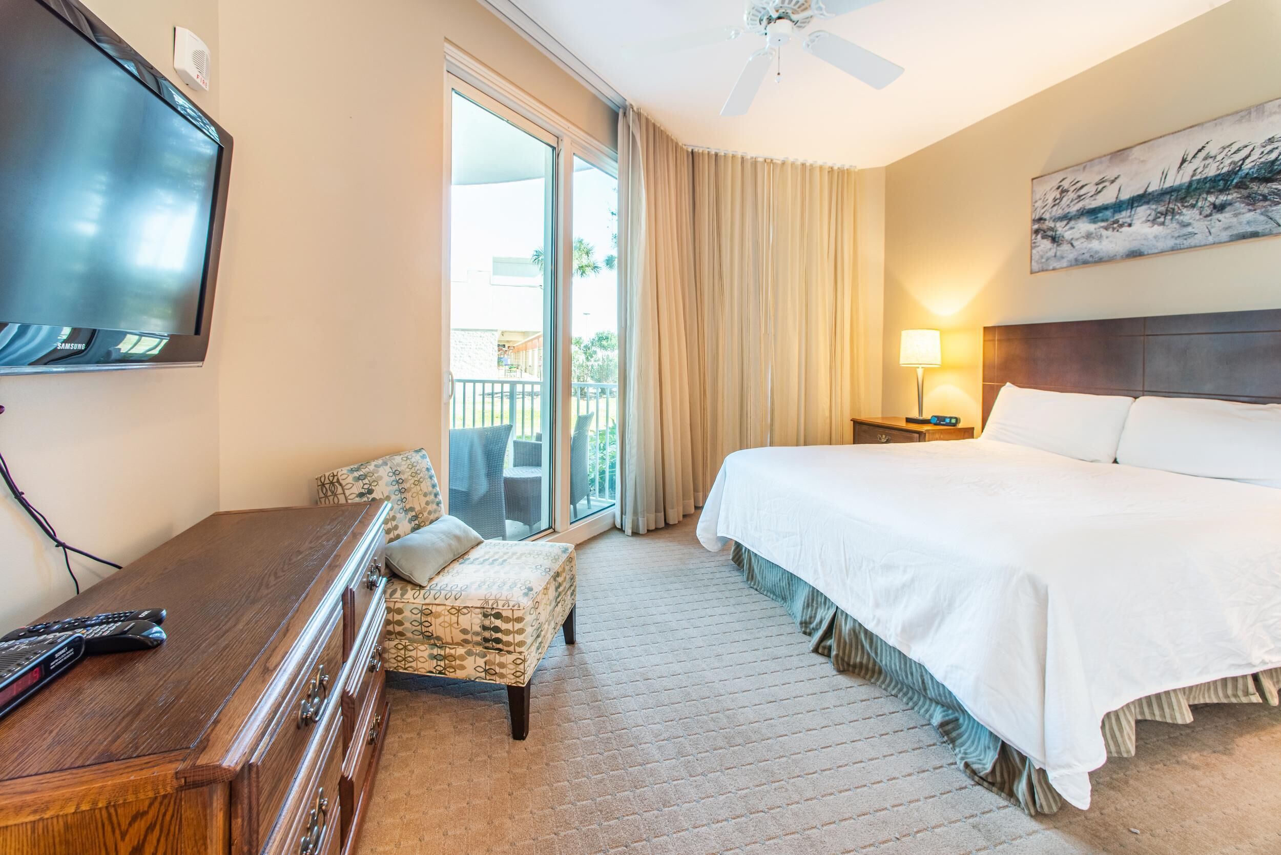 A hotel room with a large bed, a flat-screen TV, a desk with a chair, and a balcony with a view of palm trees.
