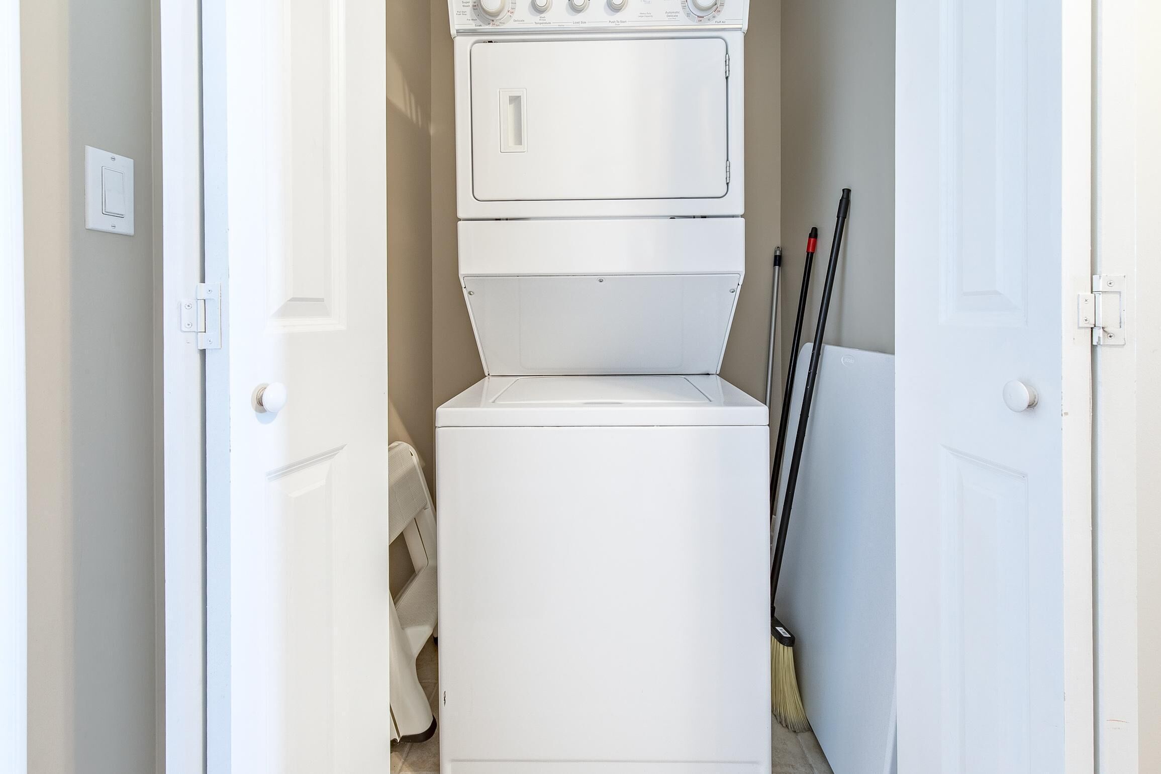 A laundry room with a stacked washer and dryer, a mop, and a broom leaning against the wall.