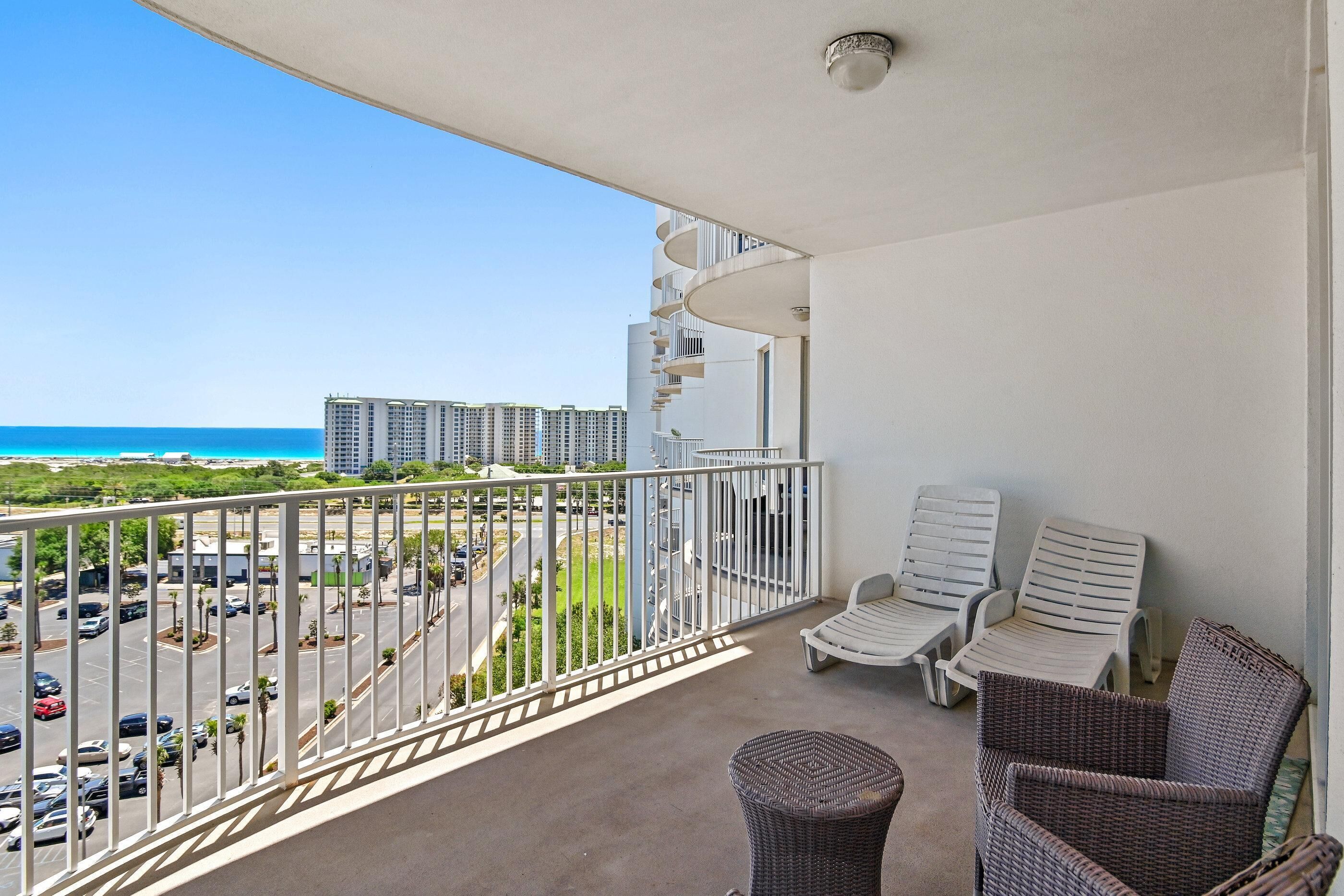 A balcony with a view of a parking lot and buildings, two wicker chairs, and a small round table.