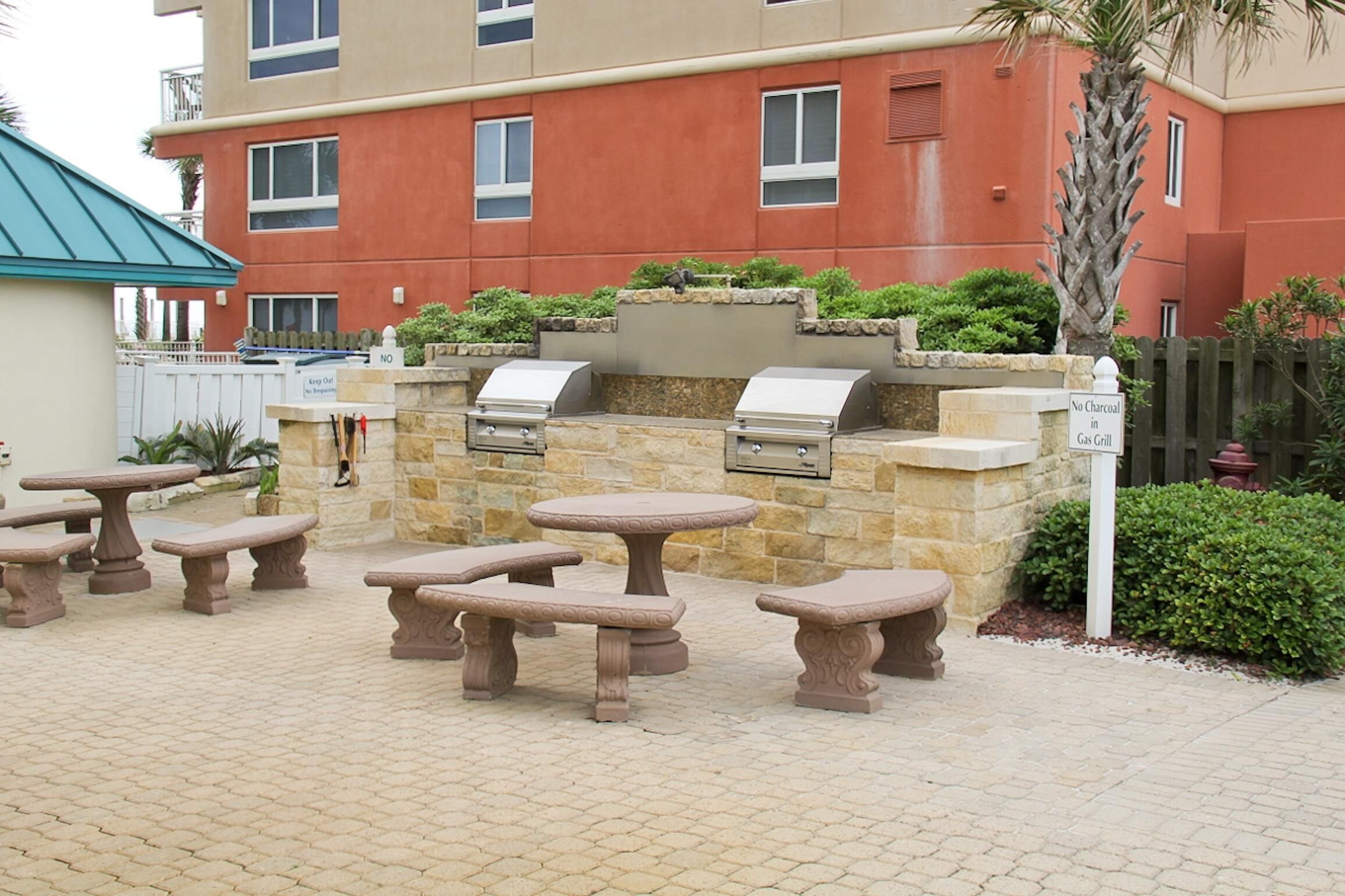 An outdoor patio with stone tables and a grill area, surrounded by a stone wall and greenery.