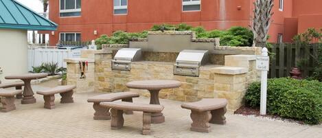An outdoor patio with stone tables and a grill area, surrounded by a stone wall and greenery.