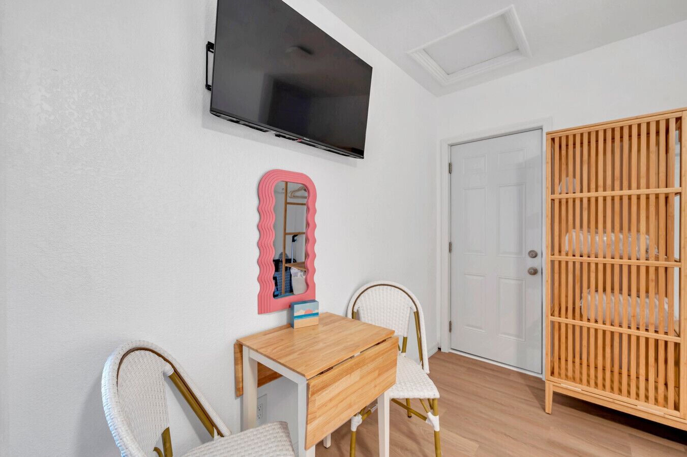 Ein Zimmer mit Schreibtisch aus Holz, zwei Stühlen, einem an der Wand befestigten Fernseher, einem Spiegel und einem Holzschrank.