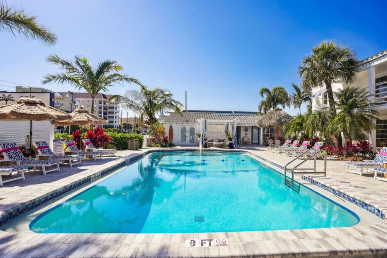 Une piscine avec des chaises longues, des palmiers et un bâtiment en arrière-plan.