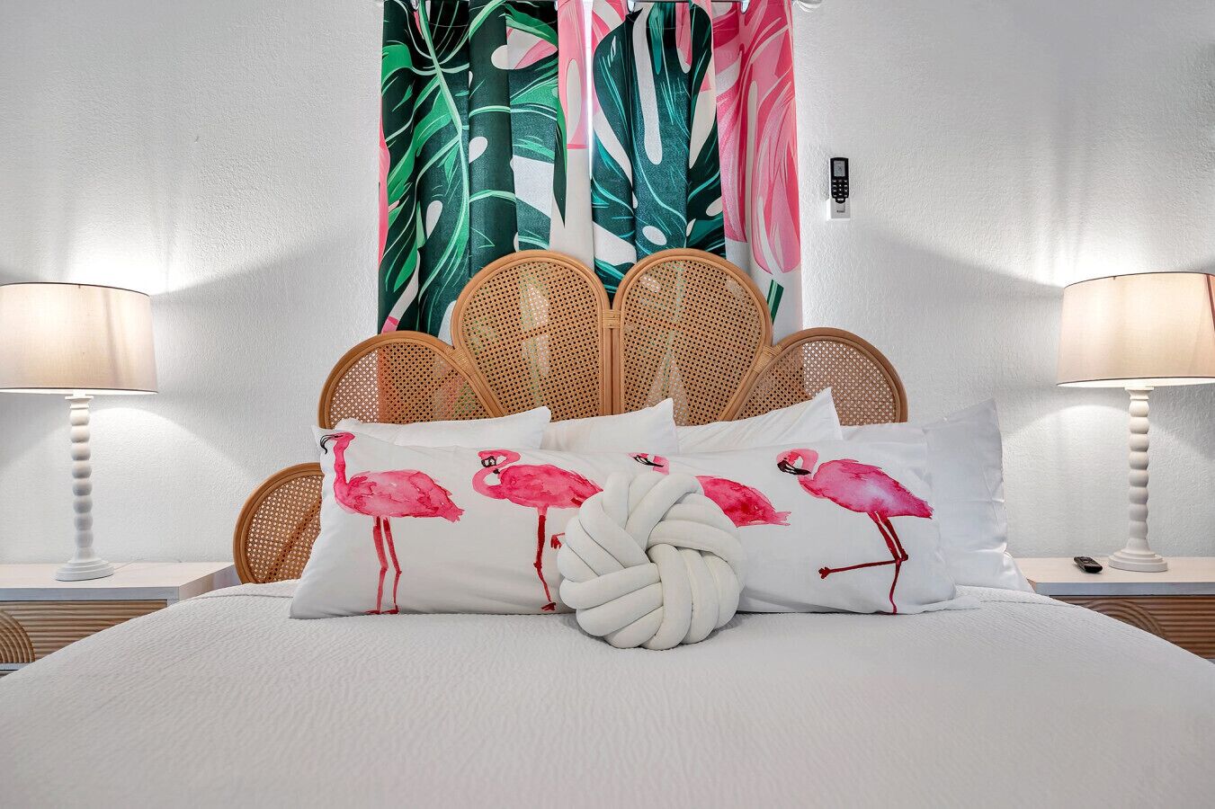 Een bed met witte lakens, een geweven hoofdbord en decoratieve kussens met flamingo's.