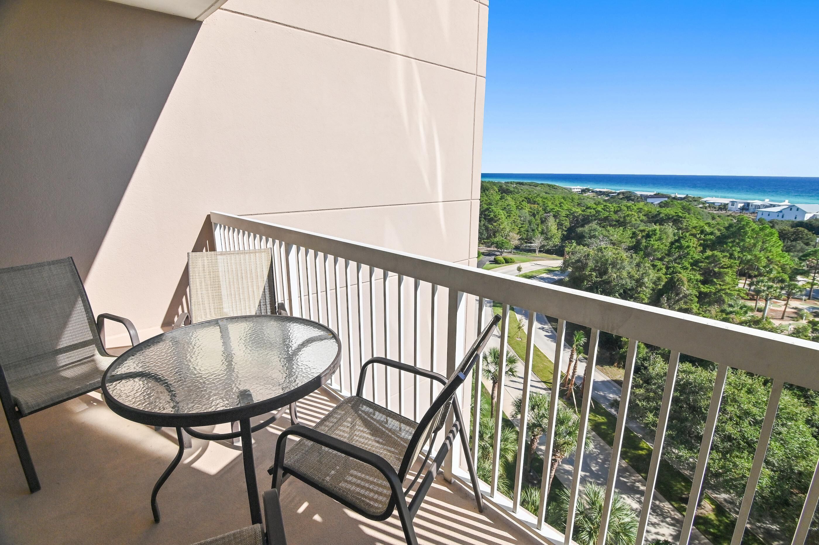 A balcony with a round table and two chairs, overlooking a coastal area with greenery and buildings.