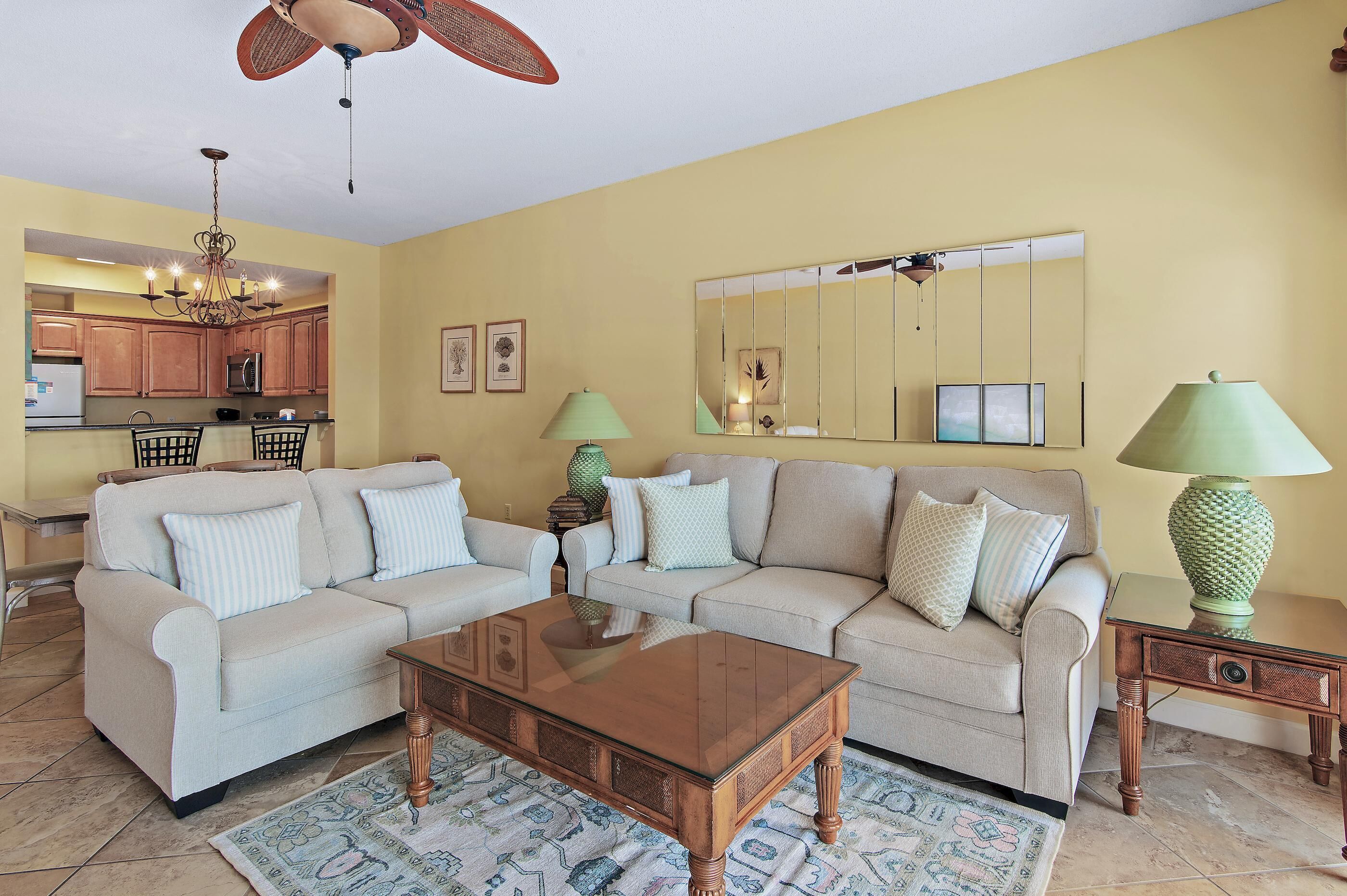 A living room with two sofas, a coffee table, a chandelier, a kitchen in the background, and a large mirror on the wall.