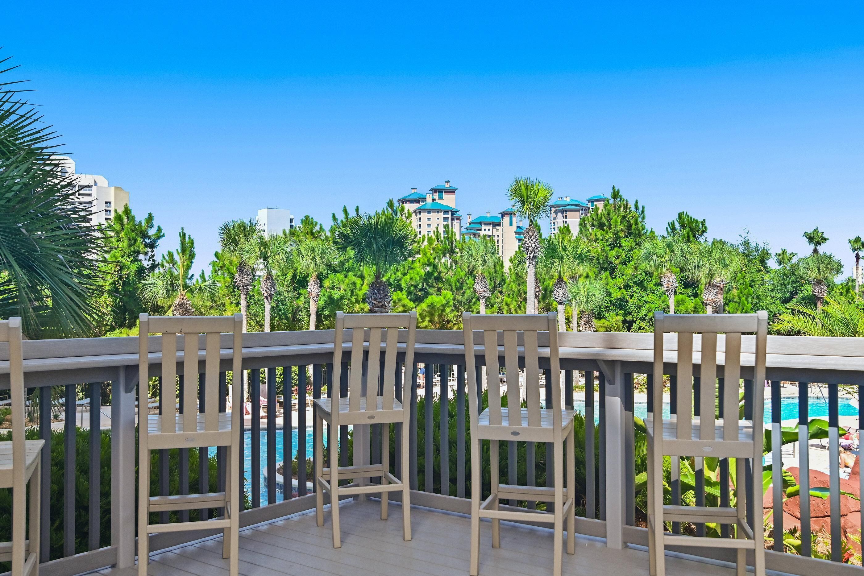 A wooden deck with chairs overlooking a pool and a view of a resort with multiple buildings and palm trees.