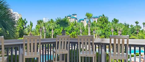 A wooden deck with chairs overlooking a pool and a view of a resort with multiple buildings and palm trees.