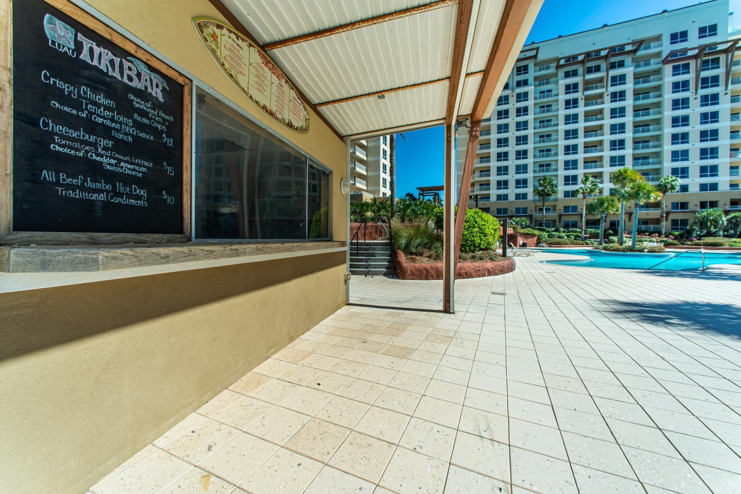Un restaurant doté d’une terrasse, d’un tableau affichant le menu, et offrant une vue sur une piscine et des immeubles de grande hauteur en arrière-plan.