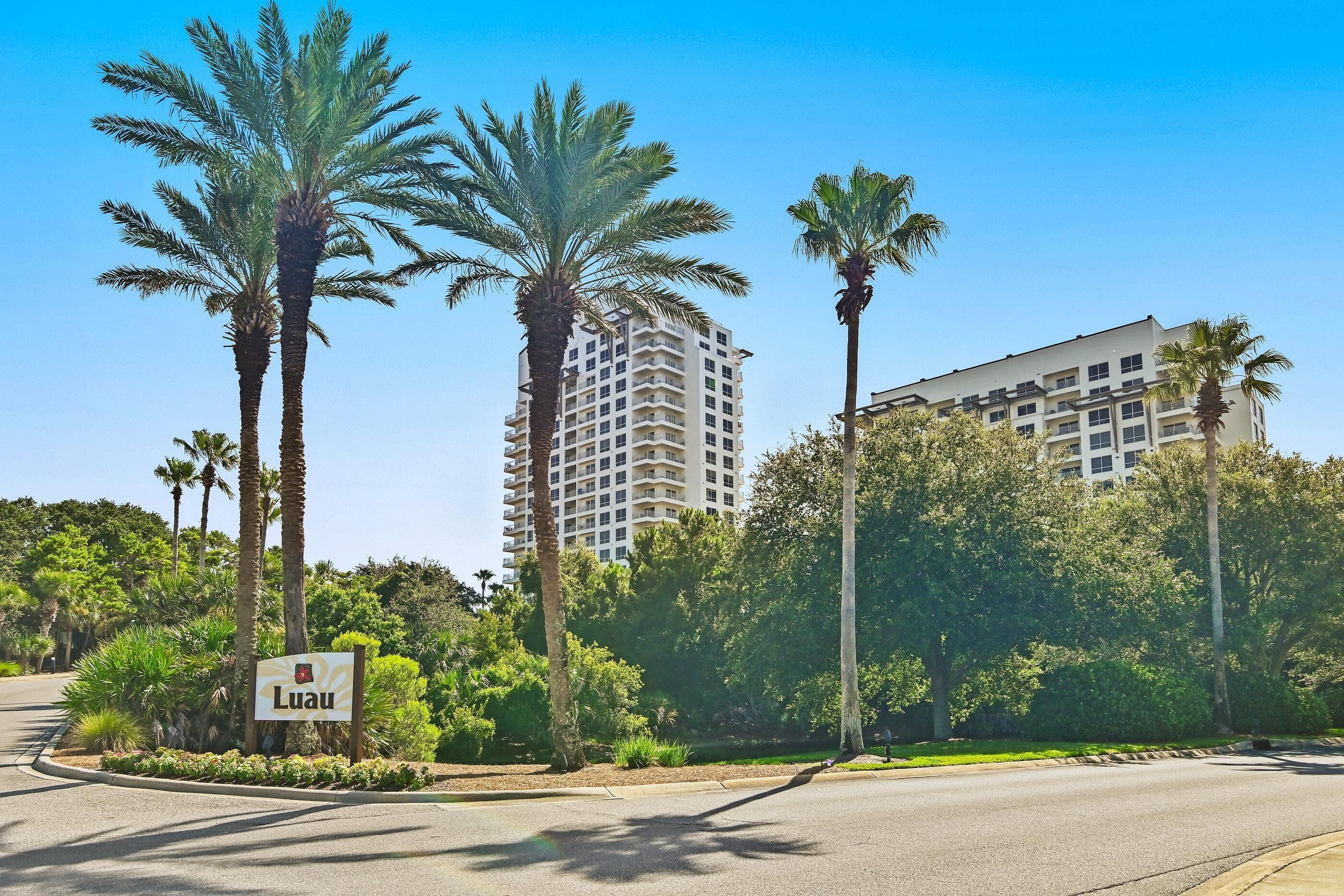 A sign with the name 'Luau' in front of a building with multiple floors and a flat roof, surrounded by palm trees and greenery.