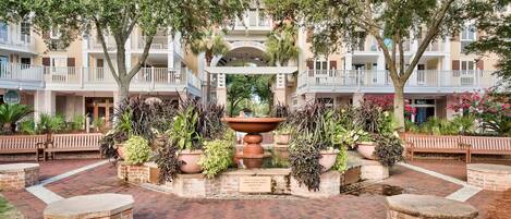A courtyard with a central fountain, surrounded by potted plants and benches, with a building featuring balconies in the background.