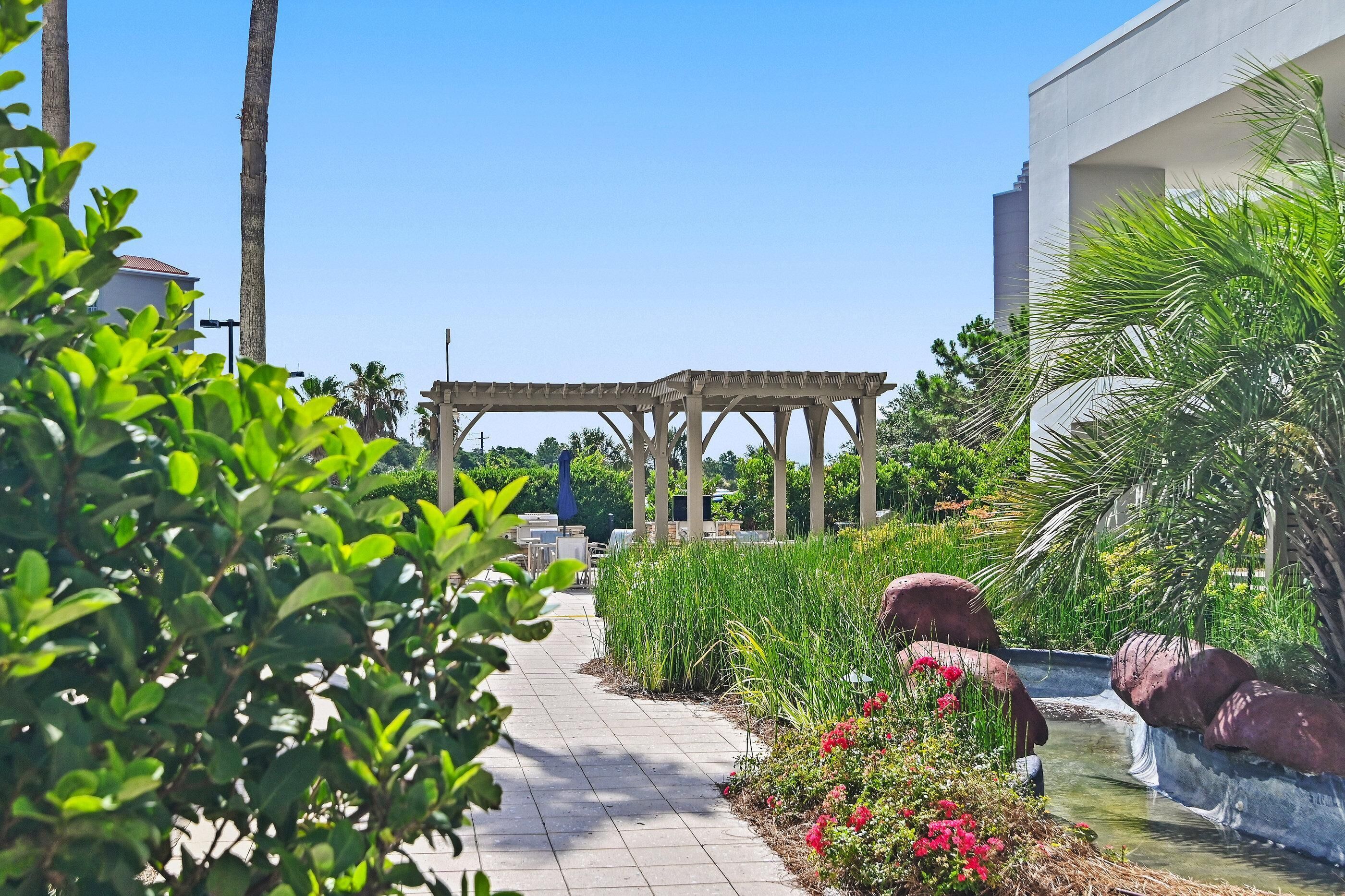 A garden pathway with a pergola, palm trees, and a water feature.