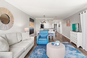 A modern living room with a sofa, a blue armchair, a round ottoman, and a dining area in the background. - Harbour Hideaway (Miramar Beach)