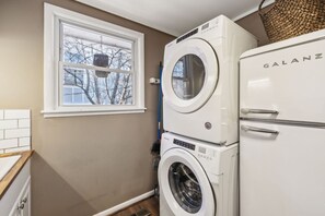 A laundry room with a washing machine and dryer stacked on top of each other, a window with a view of bare trees, and a white tiled wall.
