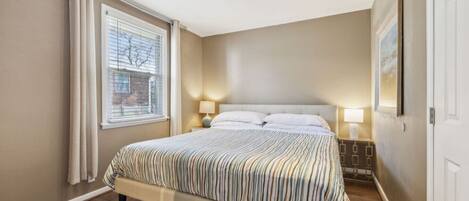 Un dormitorio con cama, dos lámparas de noche, una ventana con persianas, una puerta y un cuadro en la pared.