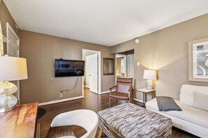 A modern hotel room with a flat-screen TV mounted on the wall, a wooden dining table, a white armchair, a patterned ottoman, and a white sofa.