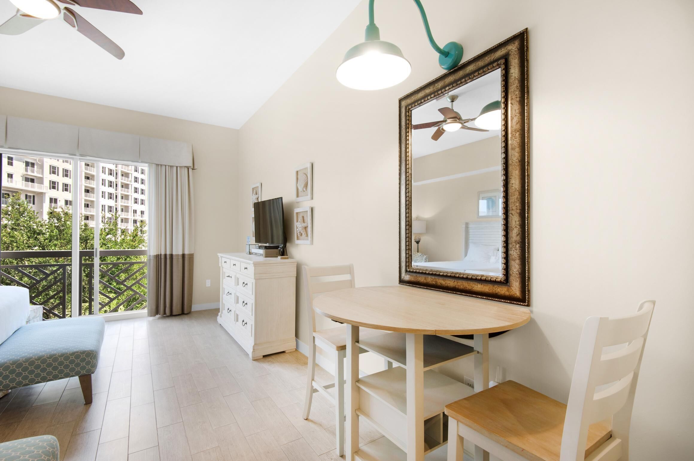 A hotel room with a balcony, a round dining table, a white dresser, and a ceiling fan.