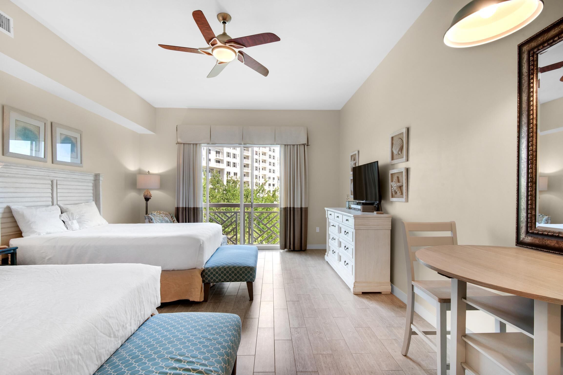 Een hotelkamer met twee bedden, een plafondventilator, een balkon met gordijnen, een televisie, een houten tafel en een dressoir.