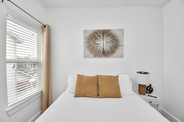 A hotel room with a bed, a window with blinds, a wall-mounted artwork, and a bedside table with a lamp. - NEW,Vibrant, 4BR house, centrally located, photo wall, Gated (Houston)
