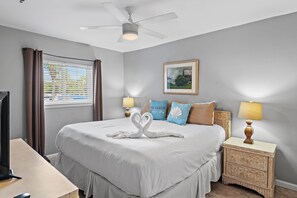 A hotel room with a bed, a television, a desk, a nightstand, a lamp, a ceiling fan, a window with curtains, and a framed picture on the wall. - Windancer 111 (Miramar Beach)