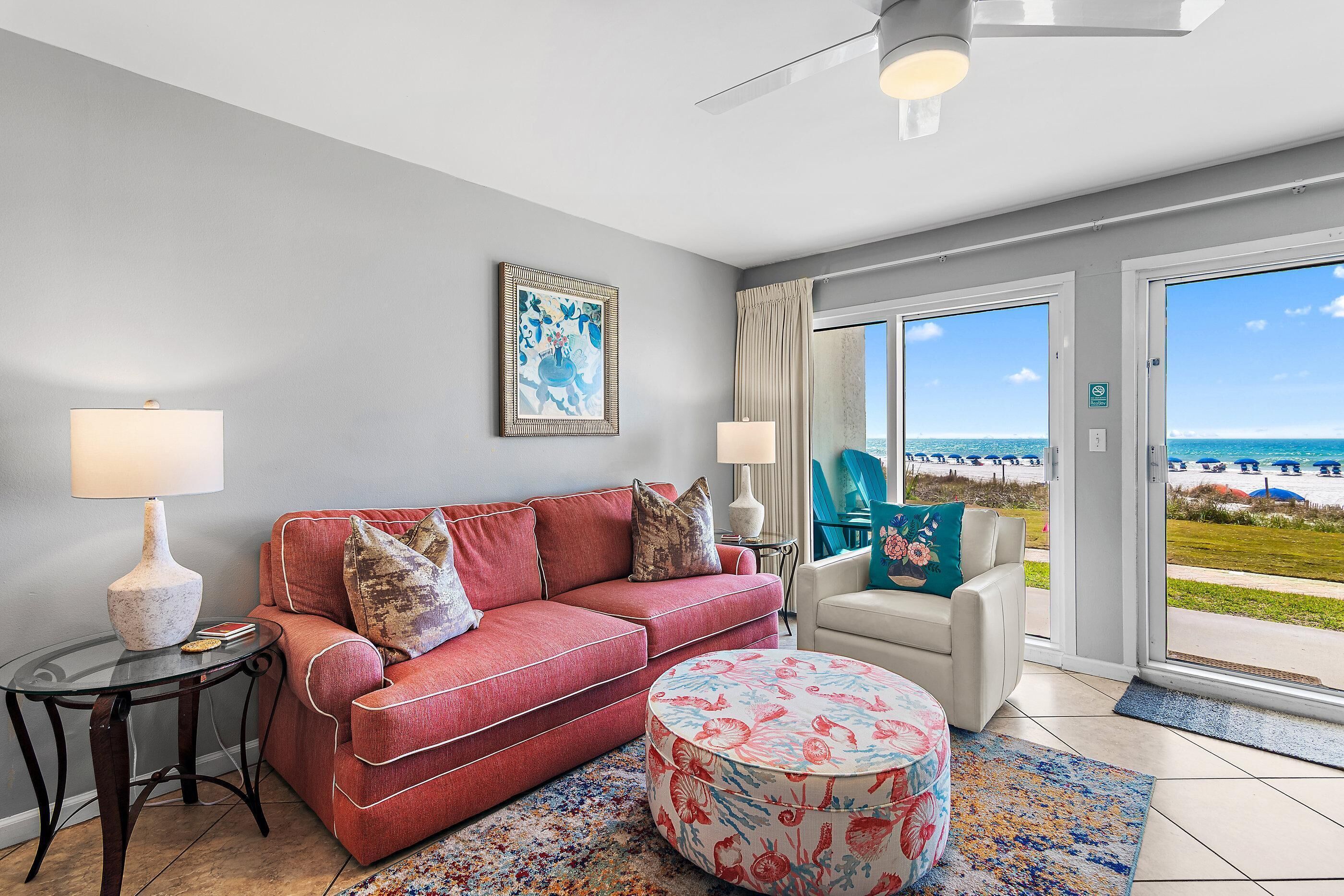 Ein Zimmer mit einem roten Sofa, einem runden Hocker, einem Glastisch, einer Lampe und Blick auf den Strand durch große Fenster.