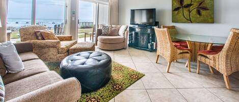 A spacious living area with a large window overlooking the beach, a flat-screen TV, and a dining table with wicker chairs.