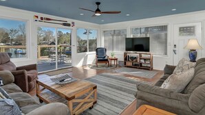 A spacious living room with a ceiling fan, large windows, a flat-screen TV, and a fireplace. - Lure & Leisure Lakehouse | Lake Murray 3BR 2BA (Prosperity)