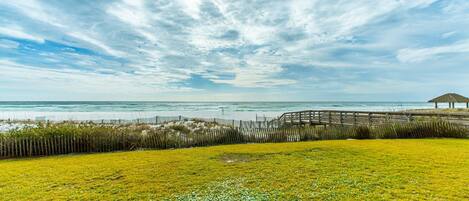 A coastal landscape with a wooden boardwalk, grassy areas, and a beach.
