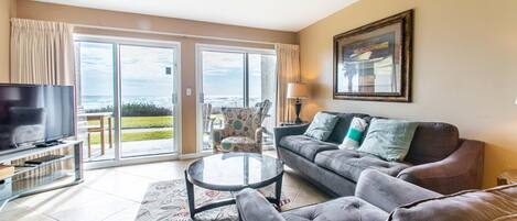 A living room with a sofa, a glass coffee table, a TV, and a view of the ocean through large sliding doors.